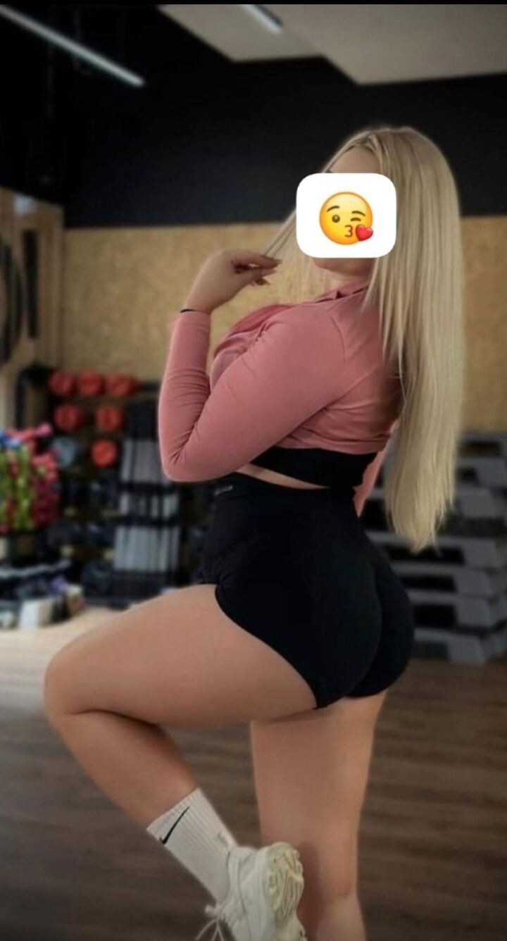 Woman posing in a gym wearing a pink long-sleeve top and black shorts, with long blonde hair and white sneakers, facing sideways.