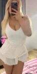 Person with long blonde hair wearing a white sleeveless top and short white skirt, taking a mirror selfie with a phone covering their face.