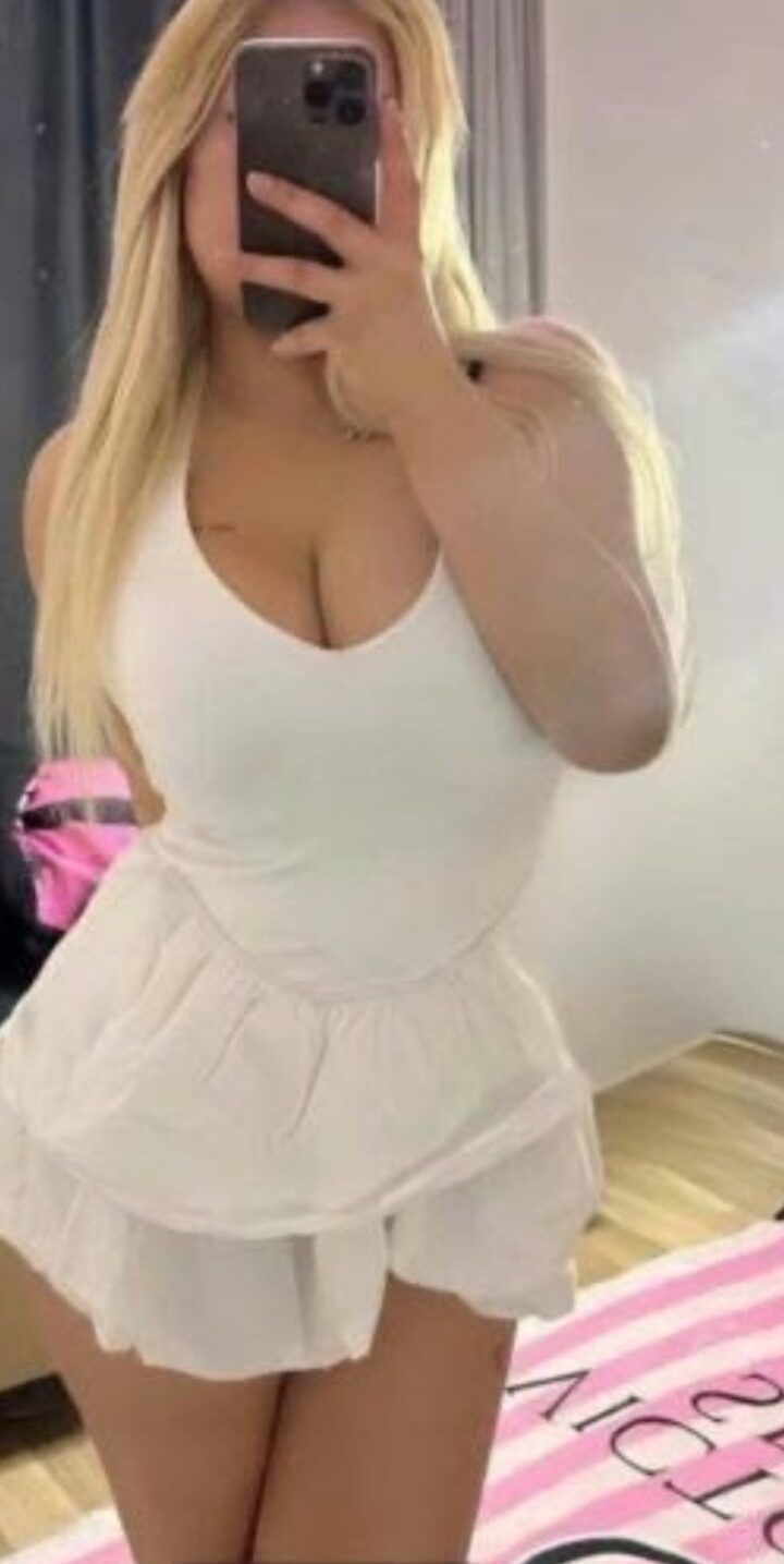 Person with long blonde hair wearing a white sleeveless top and short white skirt, taking a mirror selfie with a phone covering their face.