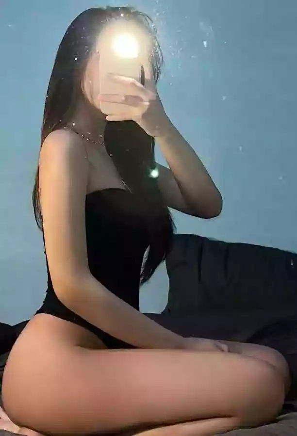 Person in a black one-piece swimsuit sits on a bed and takes a mirror selfie with a phone, face obscured by the device, blue wall in background.