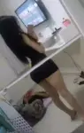 Person taking a mirror selfie in a bedroom, wearing a black crop top and shorts, with long dark hair and a phone in hand.