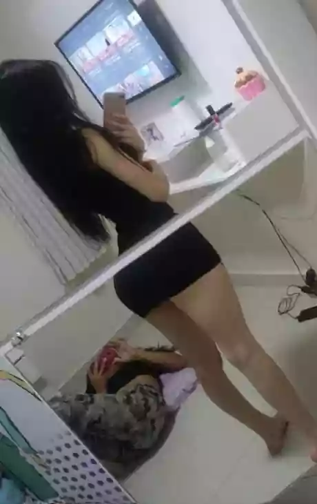 Person taking a mirror selfie in a bedroom, wearing a black crop top and shorts, with long dark hair and a phone in hand.