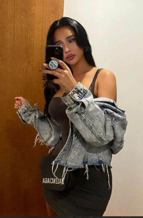 Woman posing in a mirror, wearing a gray tank dress and oversized denim jacket, holding a phone with a blue pop socket visible in the reflection.