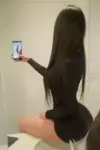 Side view of a woman with long dark hair in a black outfit, sitting and taking a selfie with a smartphone in her hand indoors.