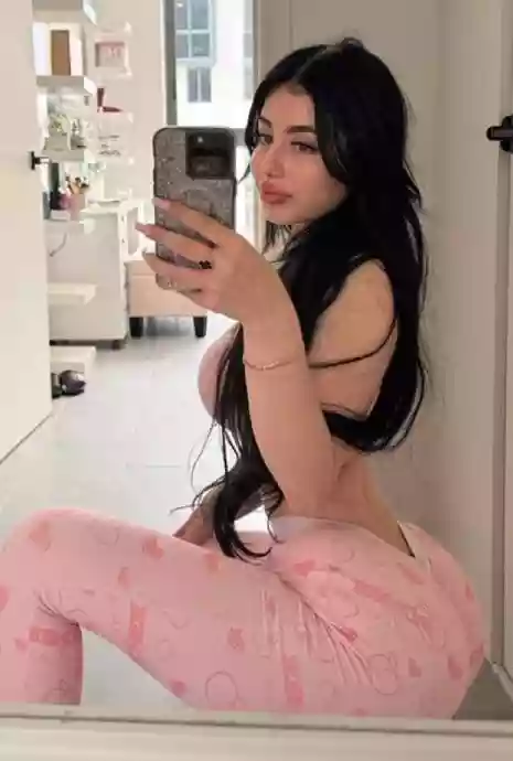 Woman with long dark hair taking a mirror selfie in a bright room, wearing pink patterned leggings.