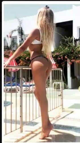 Woman in white bikini stands by pool railing, back to camera, at a sunny resort poolside.