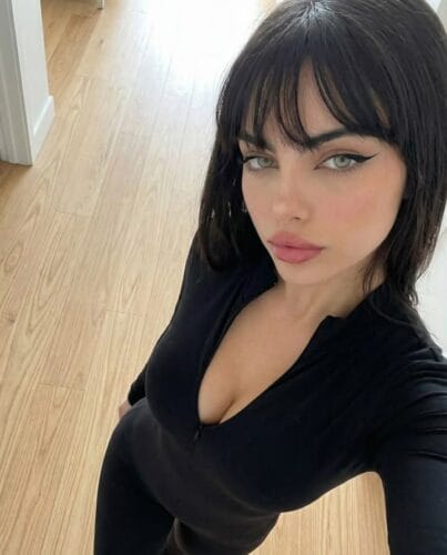 Selfie of a woman with dark hair and bangs wearing a black top, standing in a hallway with light wood floors and white walls, looking up at the camera.