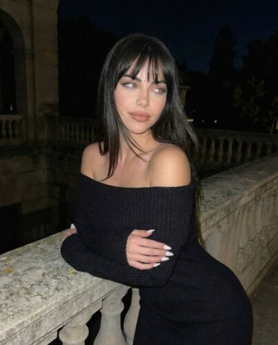 Woman with dark hair in a black off-the-shoulder sweater leaning on a stone railing at night, gazing to the side.