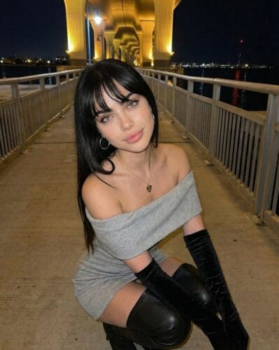 Young woman kneeling on a lit bridge at night, wearing an off-the-shoulder gray dress with long black gloves and thigh-high boots, with illuminated arches in the background.