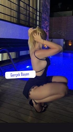 Blonde woman in a black swimsuit kneels on a wooden pool deck beside a blue-lit pool; label reads 'Gerçek Bayan'.