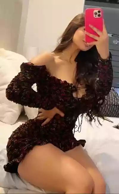 Woman posing on a bed in a dark floral off-shoulder dress, taking a mirror selfie with a pink smartphone.