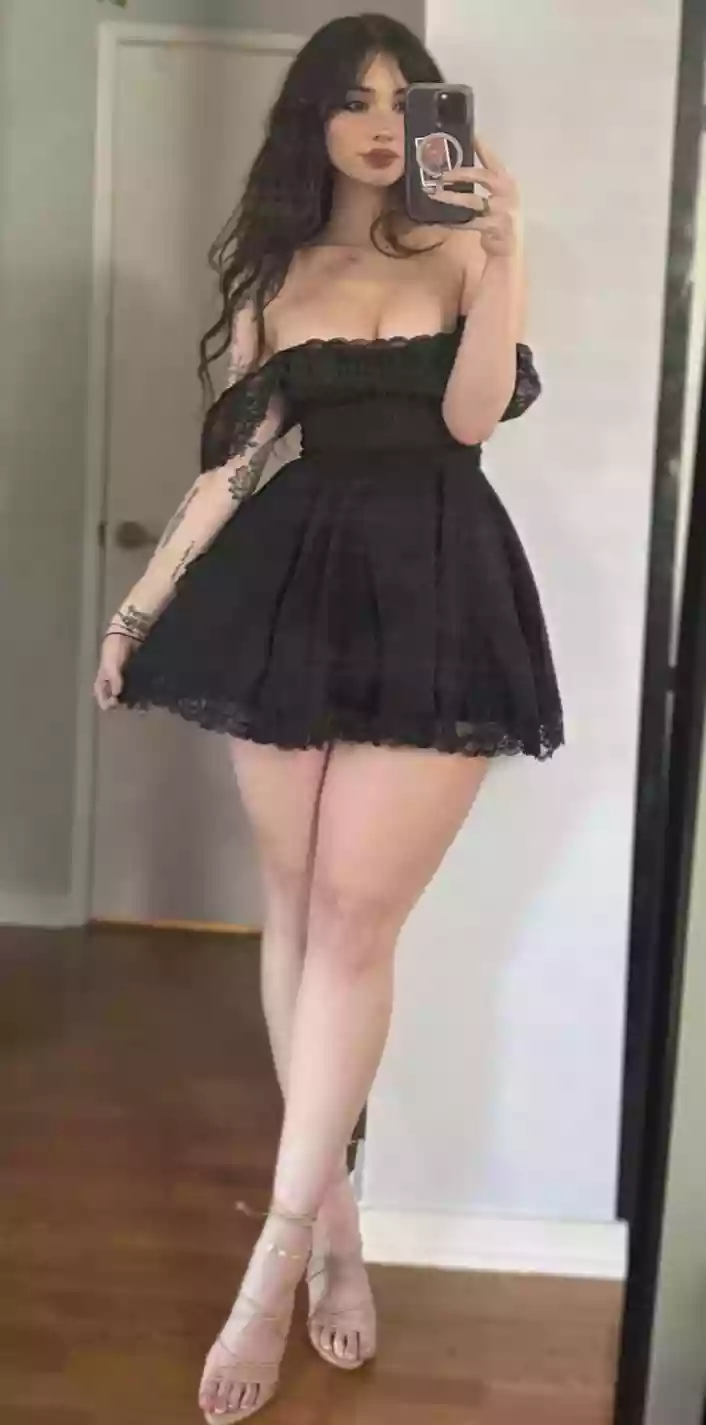 Woman wearing a short black lace off-shoulder dress, posing in front of a mirror and taking a selfie with her phone.