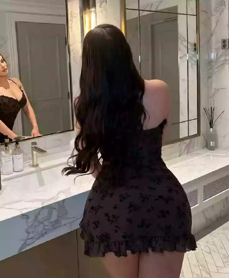 Woman with long dark hair wearing a black off-shoulder floral dress, seen from behind in a marble bathroom with mirrors.